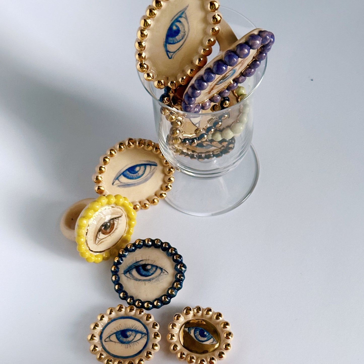 A collection of ceramic rings with various designs, including geometric patterns and eyes, displayed in a clear container.