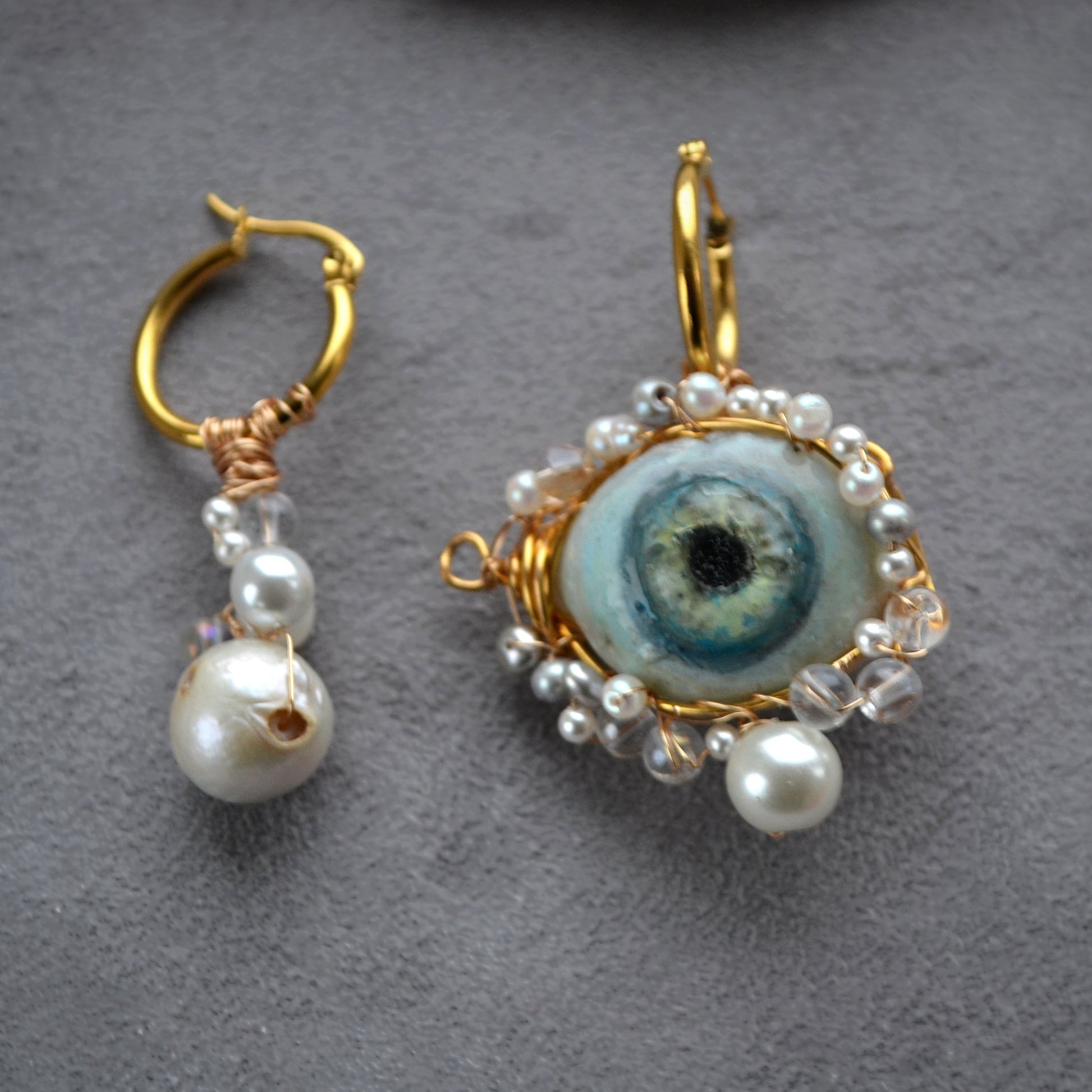Pair of earrings with gold frames, pearl accents, and a central eye design on a gray background.
