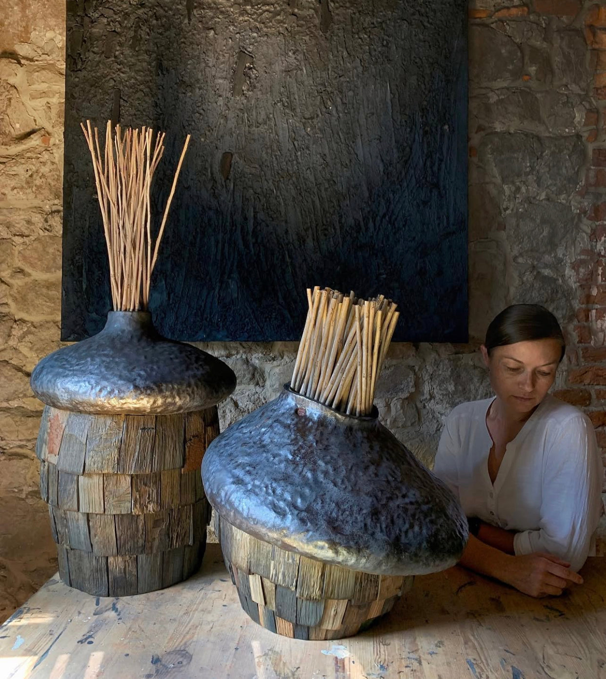 Two large decorative vases with wooden bases and black tops, one of which is being used as a incense holder, in front of a stone wall.
