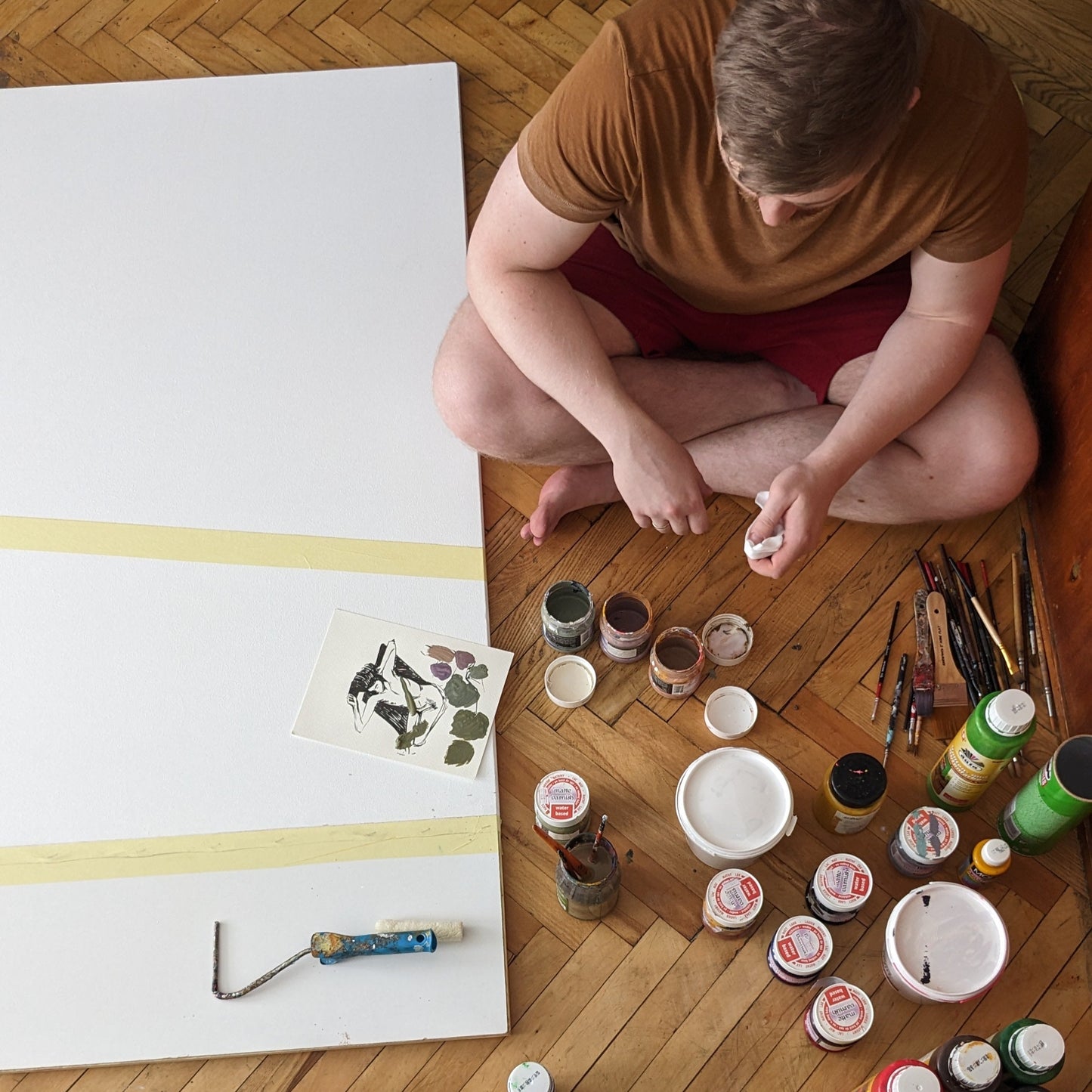 Artist sitting on a wooden floor with art supplies and a large canvas.