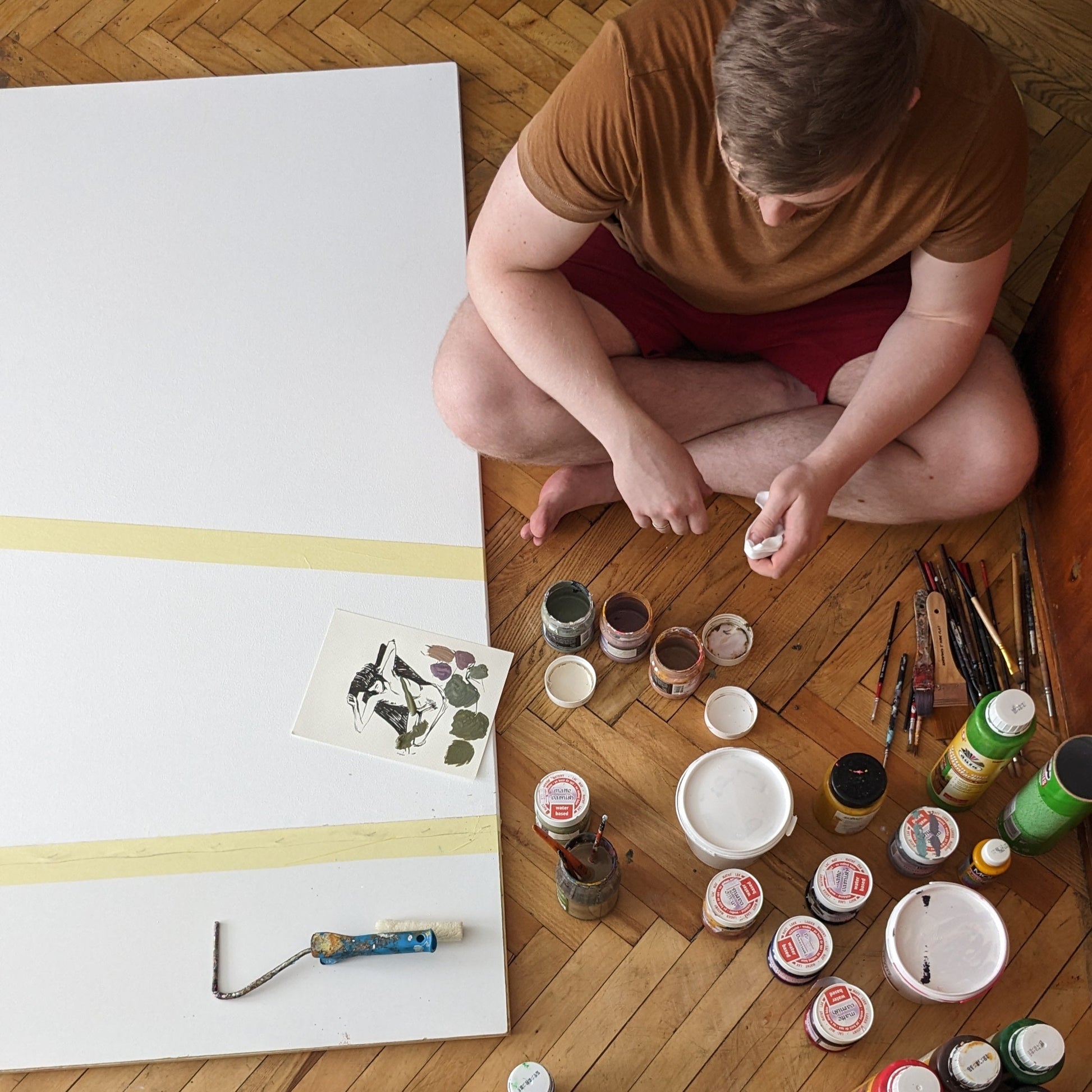 Artist sitting on a wooden floor with art supplies and a large canvas.