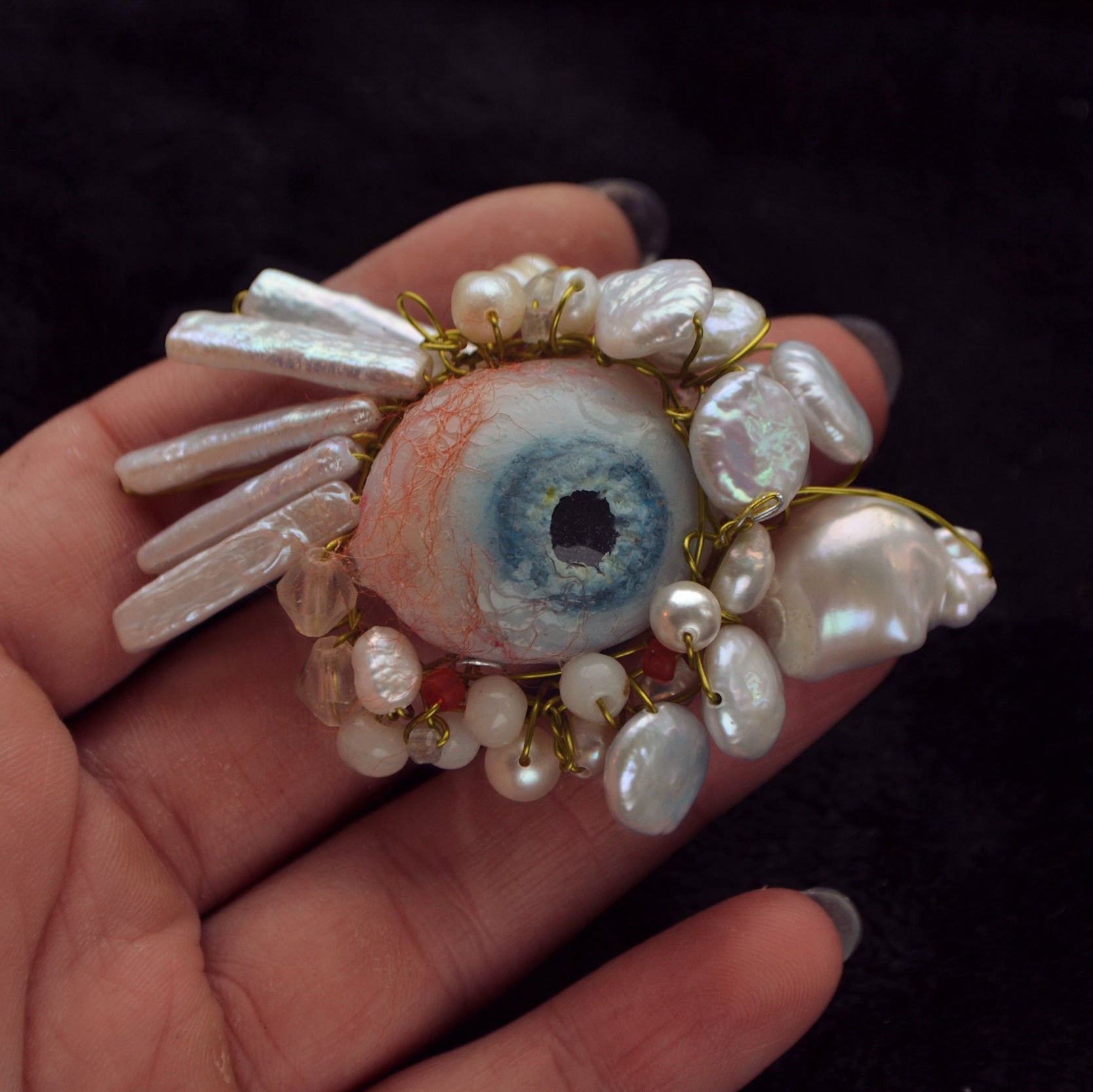 Hand holding a decorative eye-shaped object with pearls against a dark background