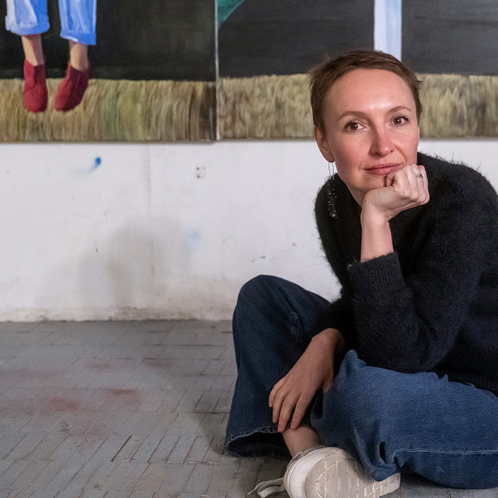 Polina Kuznetsova is sitting on the floor in an art studio with paintings on the wall.
