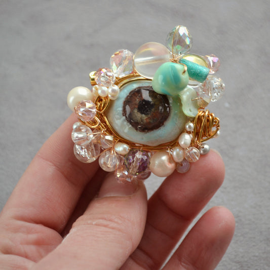 Decorative brooch with eye design held by a hand against a gray background